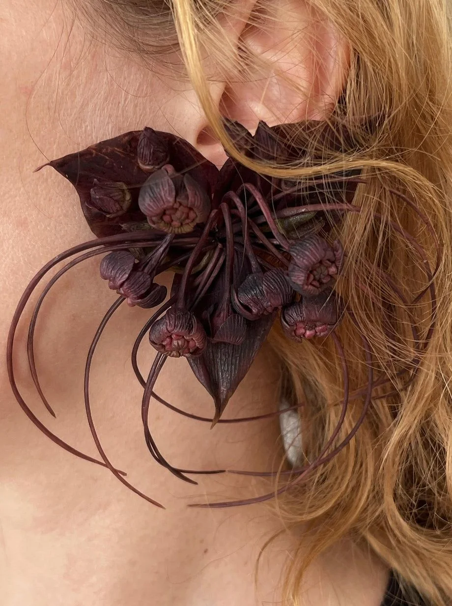A dark sculptural botanical detail beside an ear and pale hair.