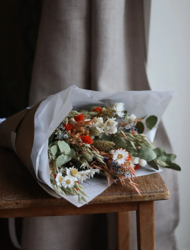A paper-wrapped botanical bouquet resting on a wooden chair beside a window.